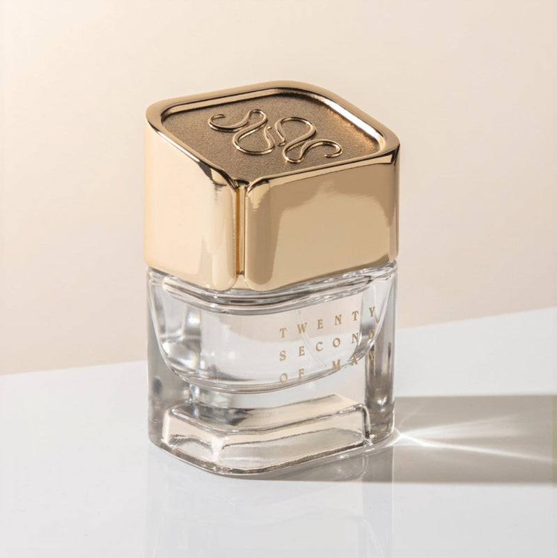 A clear glass perfume bottle with a reflective gold cap, featuring the text 'TWENTY SECOND OF MAY' on the front, set against a warm beige background on a white surface.